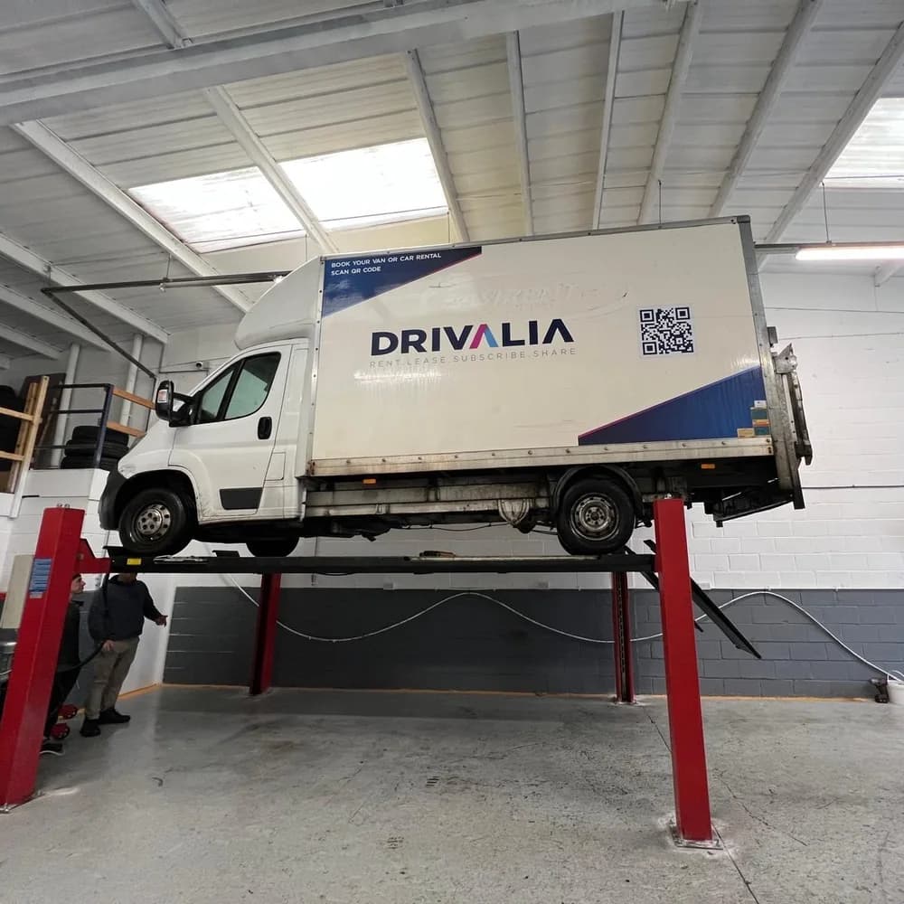 A white Drivalia Luton van raised on a four-post ramp, with two men standing beneath it.
