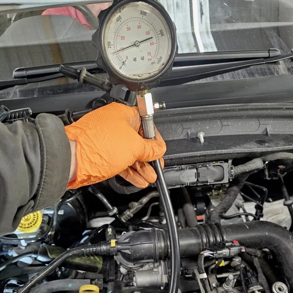 A technician in an orange glove holding a pressure gauge connected to an engine with its cover removed.