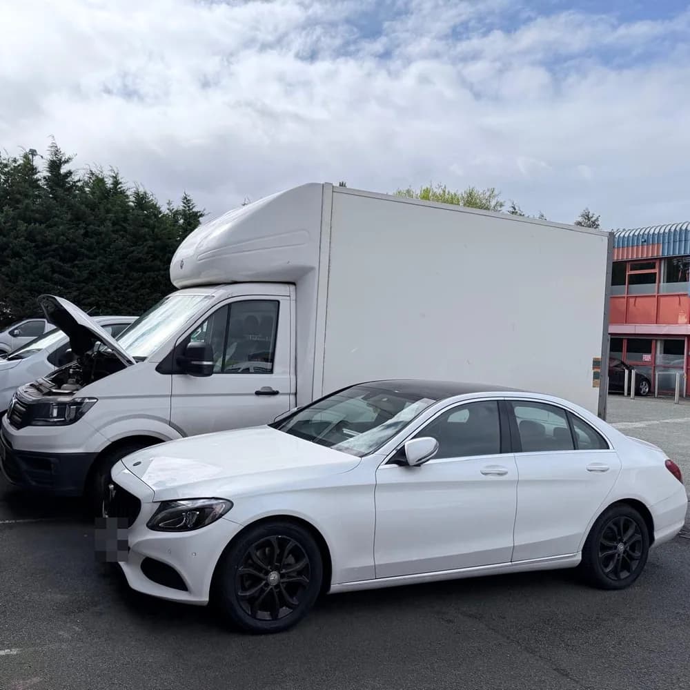 A white Mercedes-Benz saloon in the yard, beside a Volkswagen Luton van with the bonnet up.