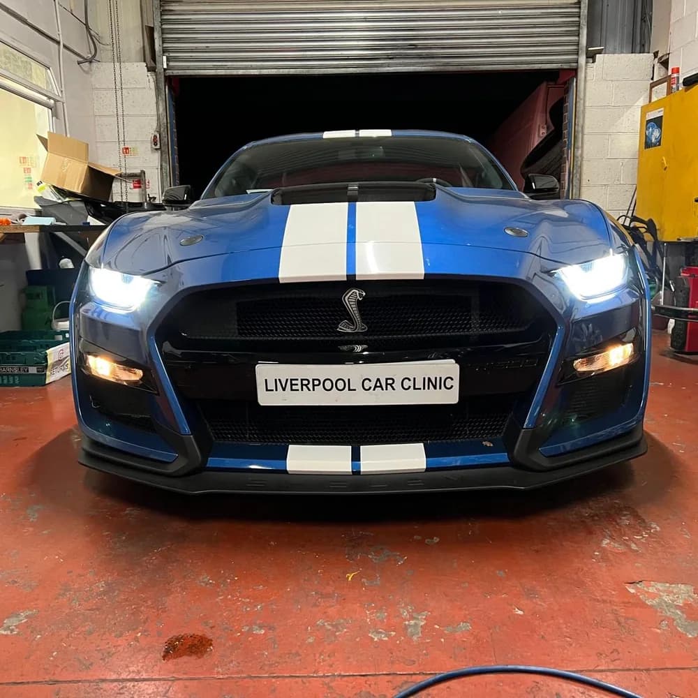 The front of a blue Shelby Mustang with its headlights on, carrying a Liverpool Car Clinic trade plate.
