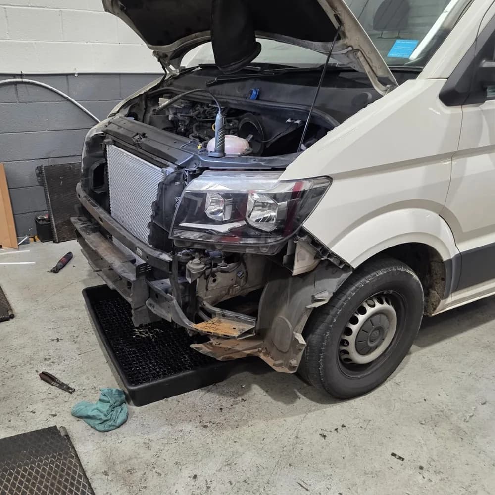 A white van in the workshop with the bonnet up and the front bumper and grille removed.