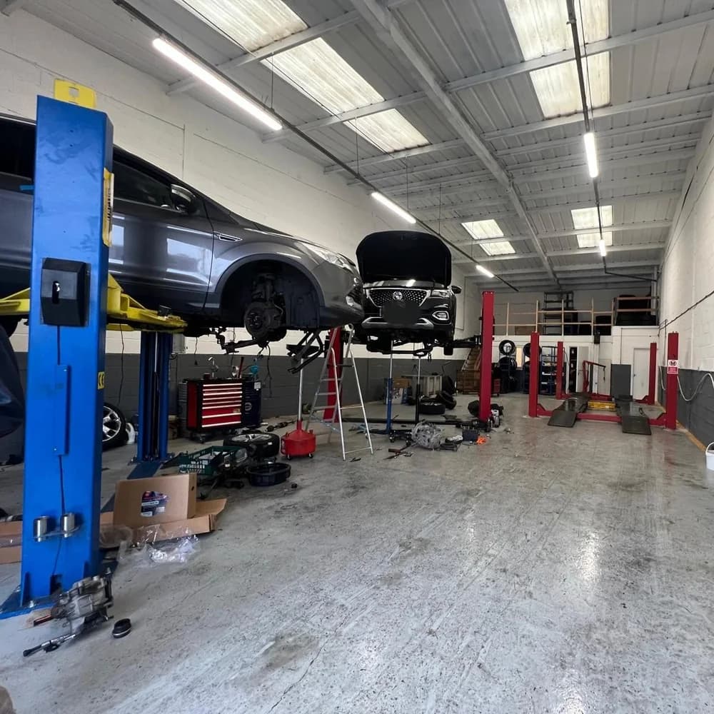 The workshop floor, with a grey SUV on a two-post lift with a front wheel off and an MG SUV raised beside it with the bonnet up.