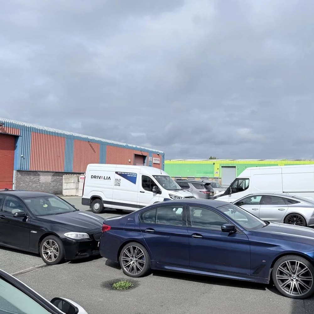 Cars and vans parked in the yard, including a Drivalia van, under a cloudy sky.