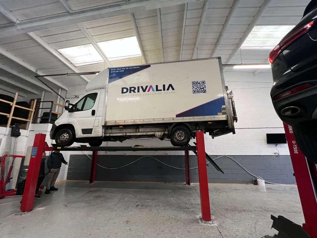 A white Drivalia Luton van raised on a four-post ramp in the Liverpool Car Clinic workshop, with two men standing beneath it.